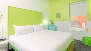 A hotel room with a bed, a nightstand, a small table, a green chair, a window, and a wall with a picture.