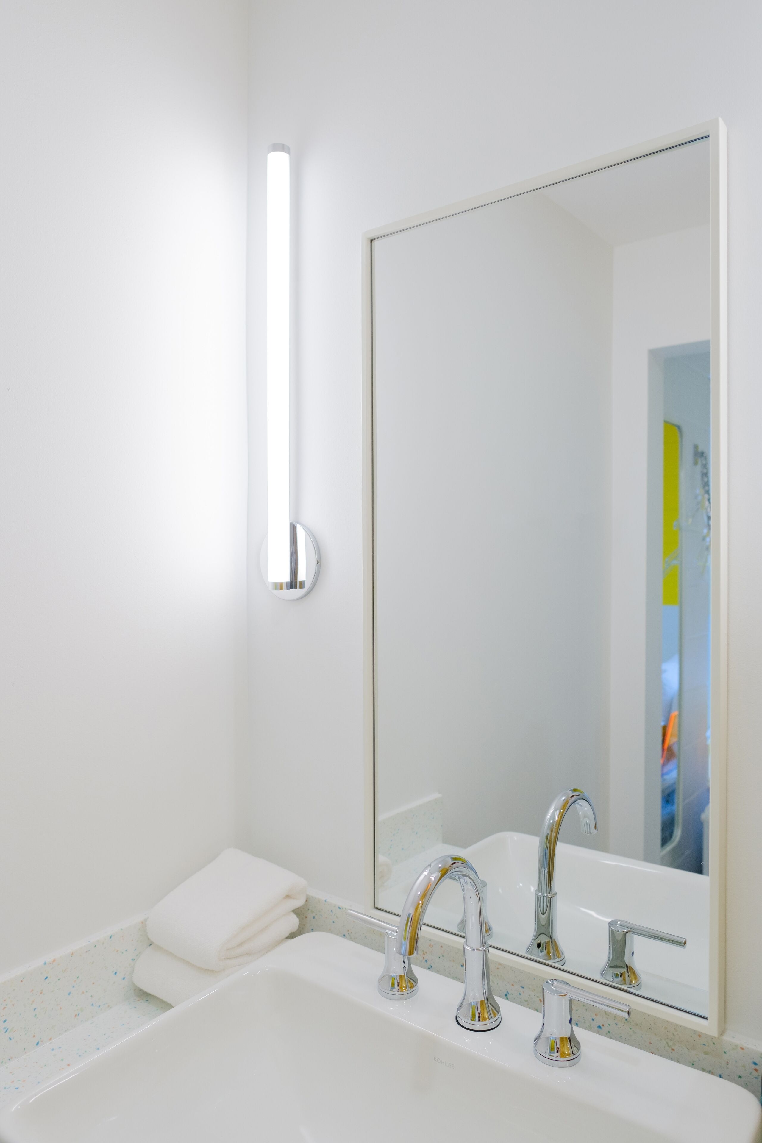 Un baño con lavamanos blanco, dos llaves, un espejo y un perchero para toallas.