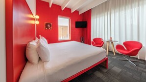 Ein Hotelzimmer mit einem Bett, roten Wänden, einem Fenster, einem Flachbildfernseher, zwei roten Sesseln und einem kleinen Tisch.