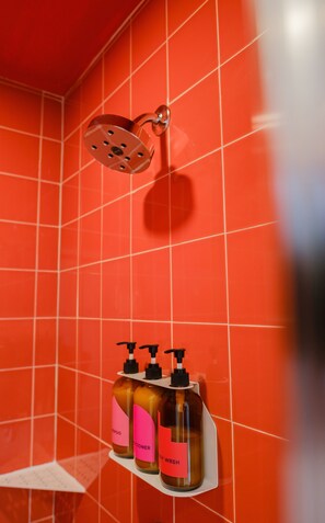 Une salle de bain avec des carreaux rouges, une pomme de douche et trois bouteilles de shampoing et d’après-shampoing sur une étagère.