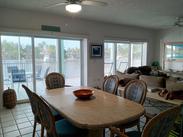 A spacious living area with a dining table, wicker chairs, a sofa, and a view of a water body through large sliding doors.