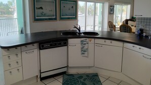 Een keuken met witte kastjes, een zwart aanrecht en een vaatwasser. Er hangen twee ingelijste foto’s aan de muur, een raam met uitzicht op palmbomen, en een plafondventilator.