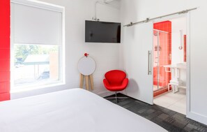 Ein modernes Hotelzimmer mit einem Bett, einem roten Sessel, einem an der Wand montierten Fernseher und einem Duschbereich mit roten Fliesen.