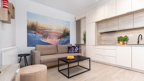 Ein modernes Wohnzimmer mit einer Couch, einem Couchtisch mit Obst, einem Gemälde an der Wand und einer Küchenzeile mit Schränken und Spüle.