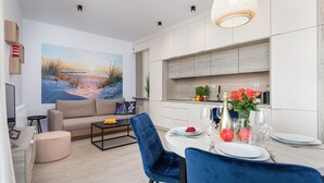 Ein modernes Wohnzimmer mit einem beigefarbenen Sofa, einem runden Esstisch für zwei Personen und einem großen Gemälde mit Strandmotiv an der Wand.