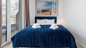 Ein Schlafzimmer mit großem Fenster, einem Bett mit blauer Bettdecke und Blick auf einen Strand.