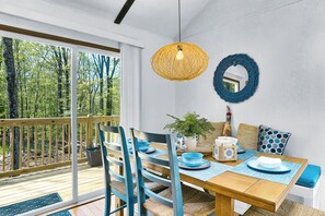 A dining area with a wooden table, blue chairs, a hanging light fixture, and a view of trees through large windows.