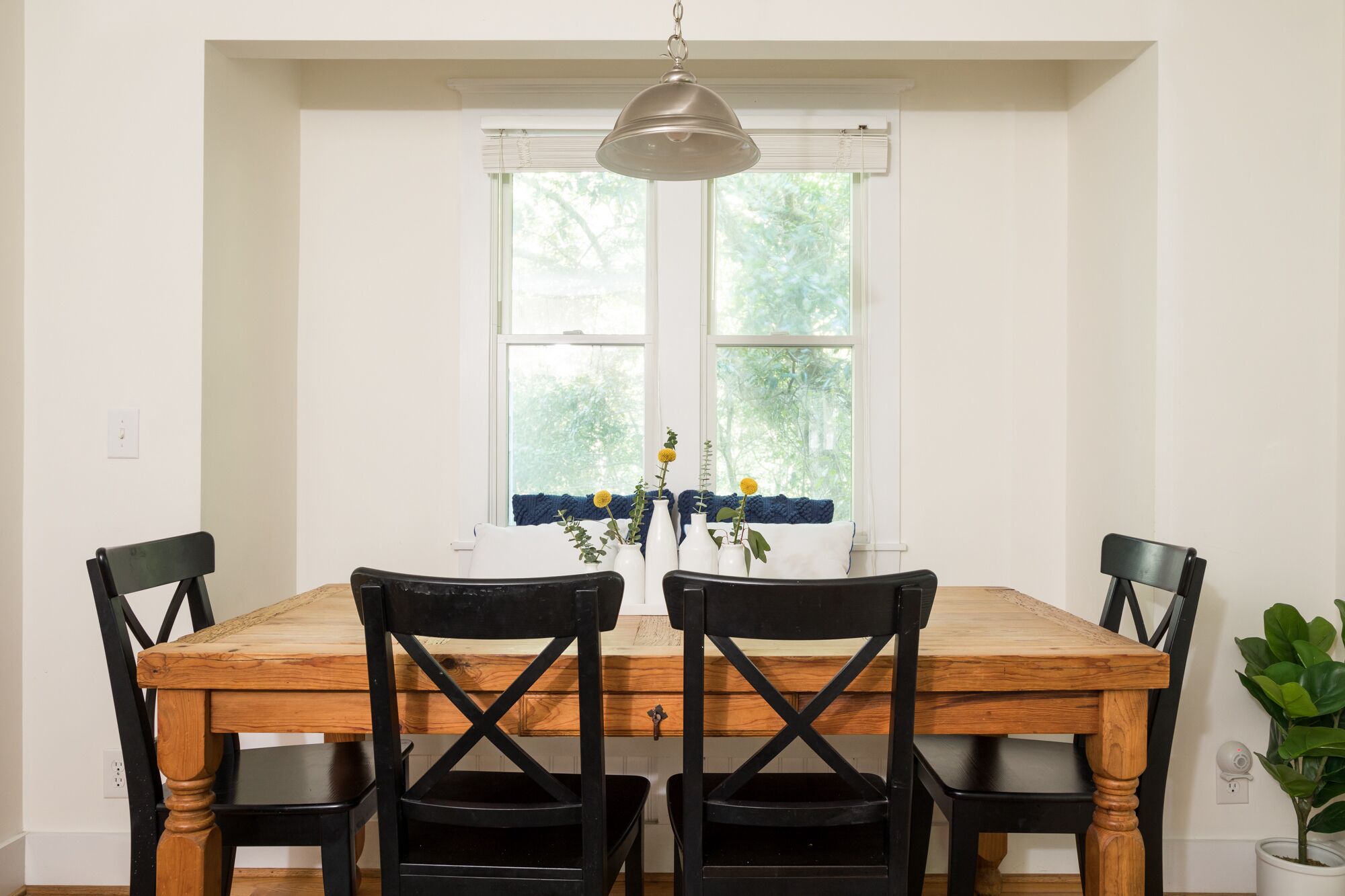 A dining table with four chairs, a hanging light fixture, and a window with a view of trees.