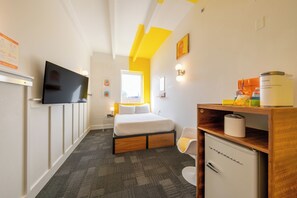 A hotel room with a bed, a television, a desk with a refrigerator, and a chair. - The Gallatin (Standard Queen Room 314) Colorful Boutique Hotel (Nashville)