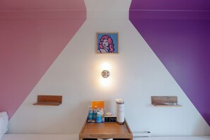 Une chambre avec un lit, une table de chevet avec une horloge, un tableau encadré et une étagère fixée au mur, sur laquelle se trouvent une bouteille d’eau et une boîte.