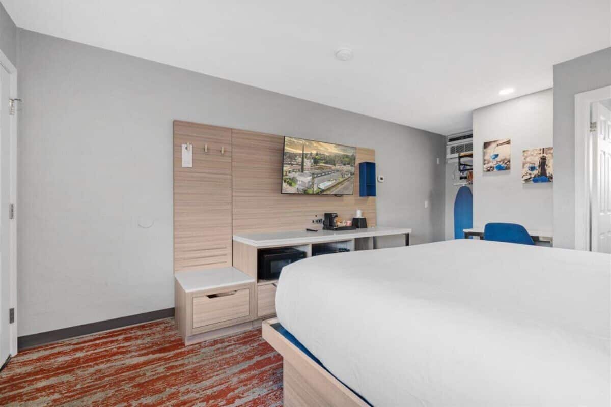 A modern hotel room with a bed, a desk, and a wardrobe. There is a painting on the wall and a small table with a lamp.