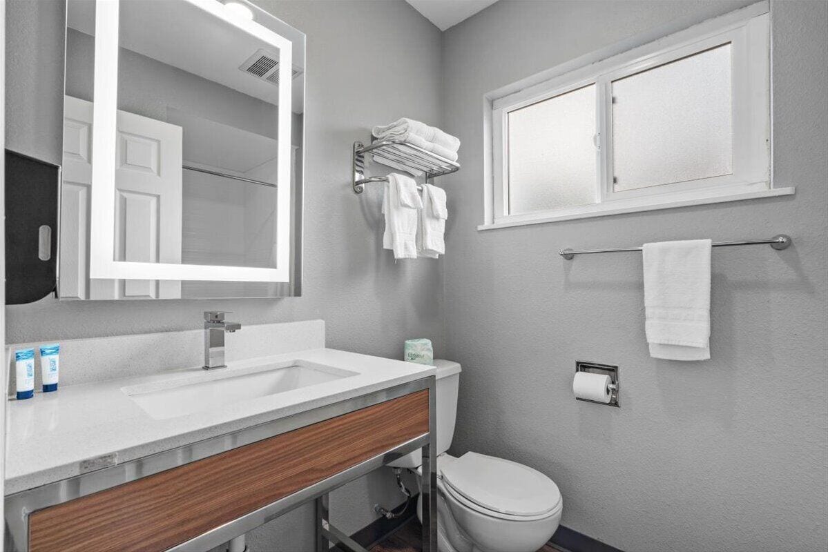 Une salle de bain avec un lavabo blanc, un miroir, un porte-serviettes, des toilettes et une fenêtre.