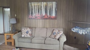 A living room with a beige sofa, patterned cushions, a glass coffee table, and a painting of trees and flowers on the wall. - Beautiful cabin on the Manistee River (Mesick)