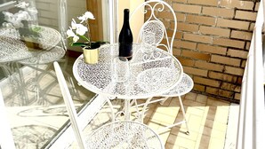Ein weißer Metalltisch mit Stühlen, einer Weinflasche und einer kleinen Pflanze auf einer gefliesten Terrasse, im Hintergrund eine Backsteinmauer.