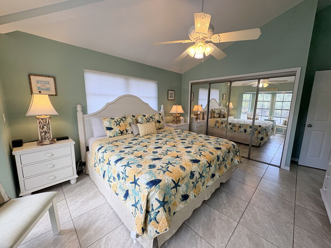 A bedroom with a large bed, a ceiling fan, a nightstand, a chair, and a mirror.