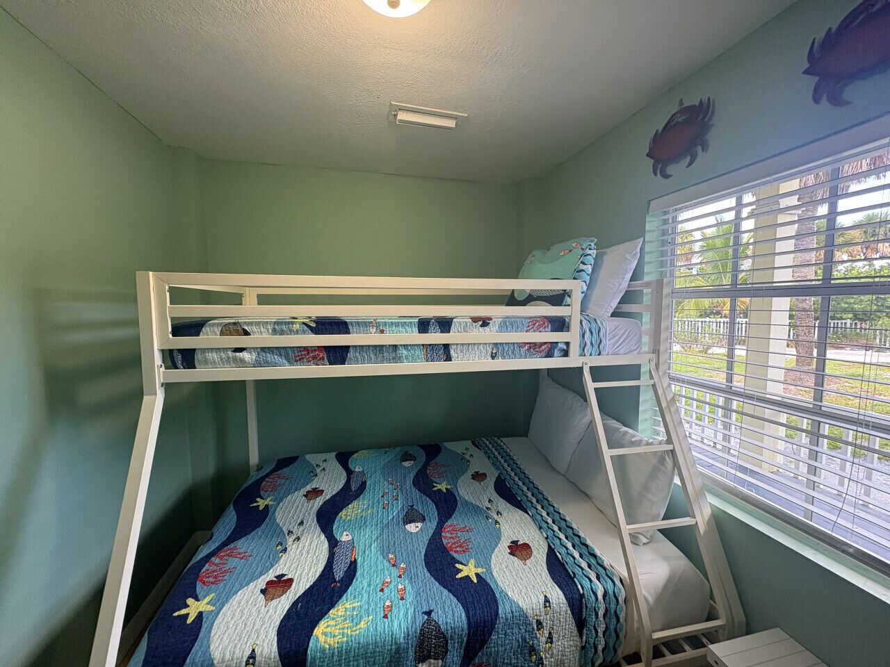 A bunk bed room with a patterned bedspread, a window with blinds, and a ceiling light.