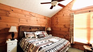 Une chambre dans une cabane en bois, avec un lit, deux tables de chevet, un ventilateur de plafond et une fenêtre avec des rideaux.