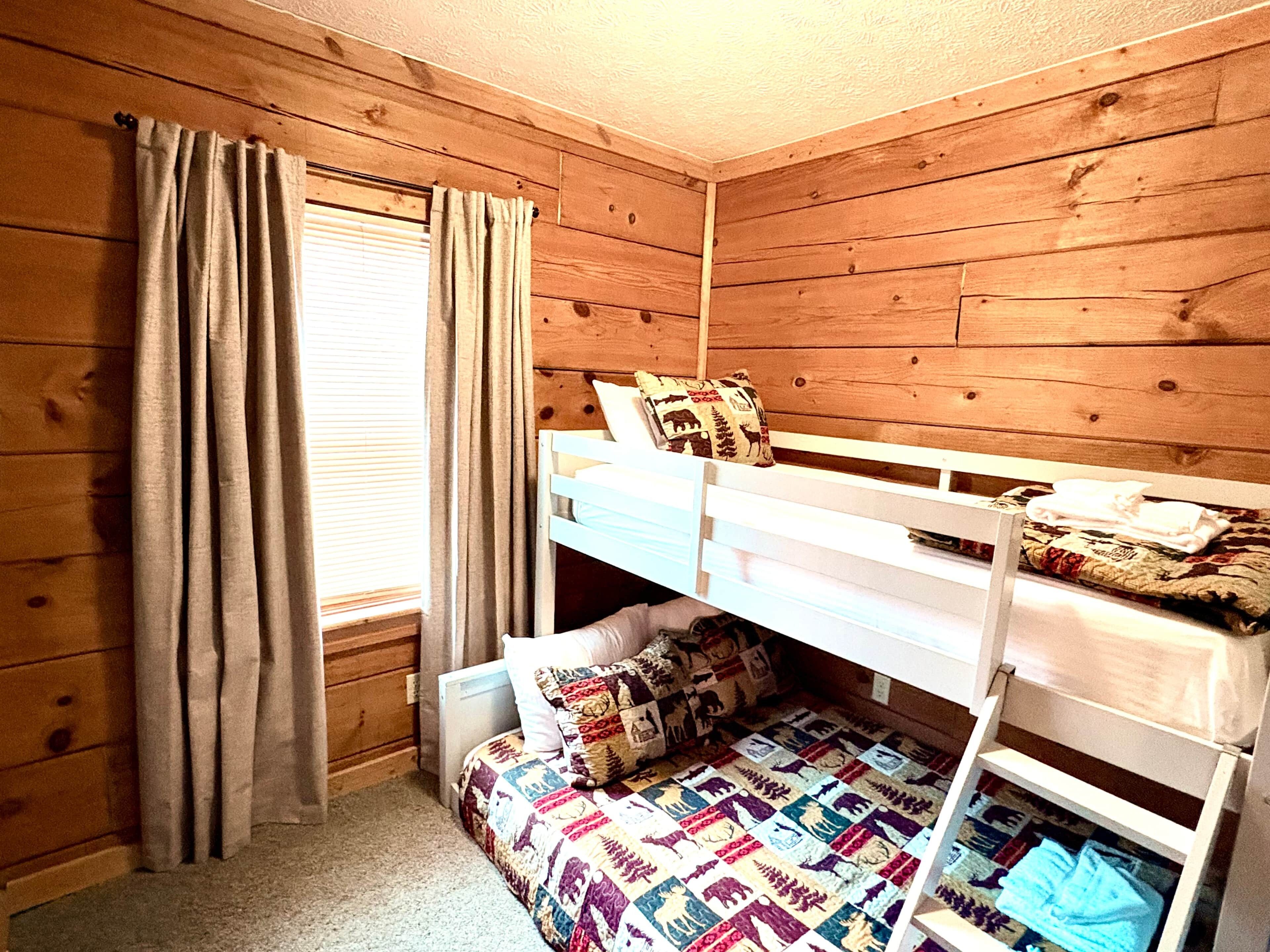 A bunk bed room with wooden walls, a window with curtains, and a quilted bedspread.