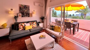 A modern living room with a grey sofa, colorful cushions, a white coffee table, and a dining area with wicker furniture and orange umbrellas. - Arma O DE P RA Twins 2 by Homing (Lagoa)