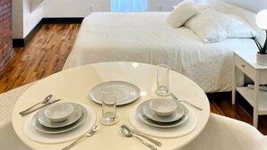 A neatly arranged dining table with plates, cutlery, and glasses, set in a room with a bed and bedside table in the background.