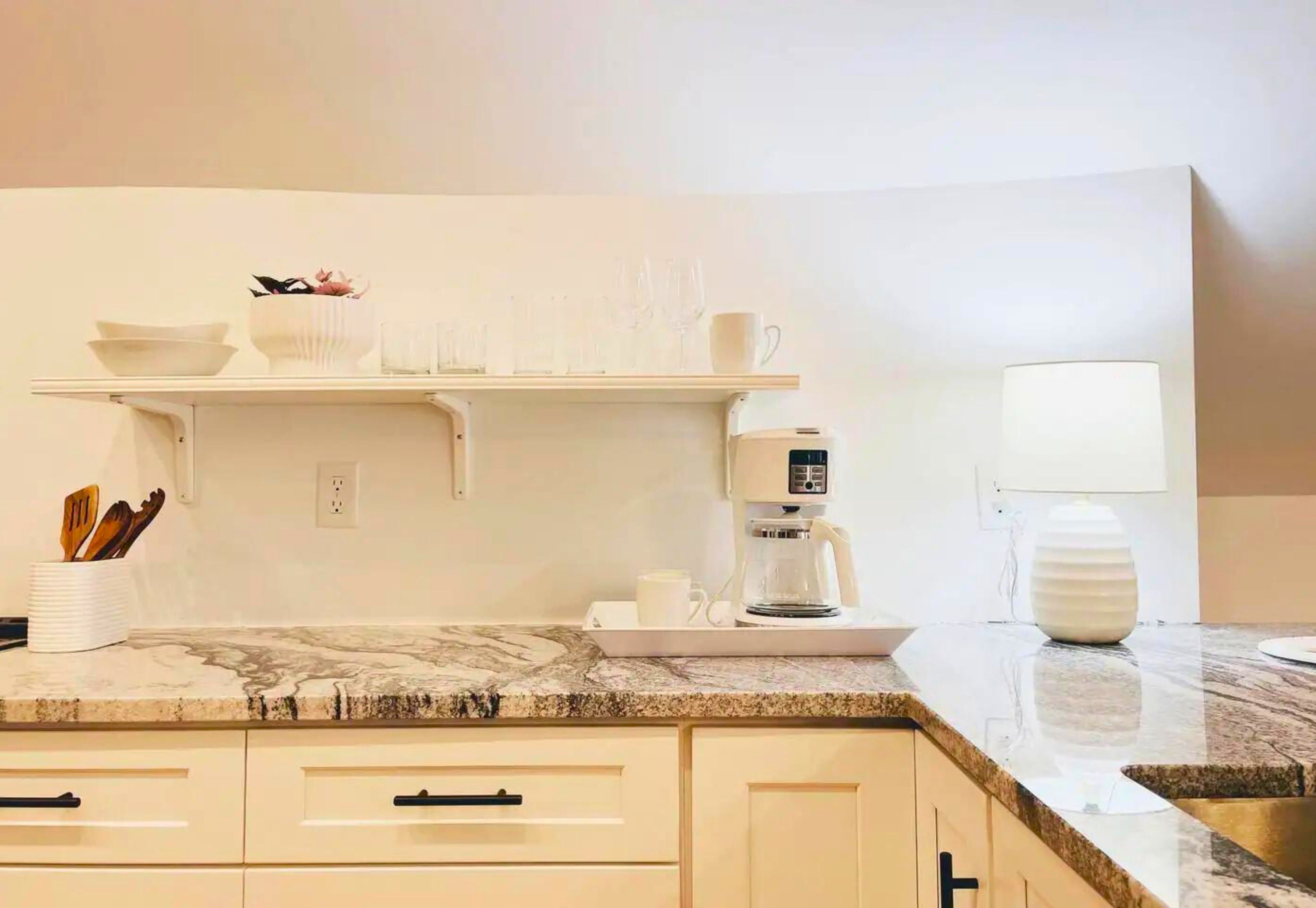 A modern kitchen with white cabinets, a marble countertop, a coffee maker, and a white vase.