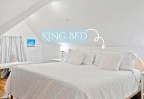 Ein Kingsize-Bett mit weißer Steppdecke, ein Nachttisch mit Lampe und Blick zum Himmel durch ein Fenster mit weißen Vorhängen.