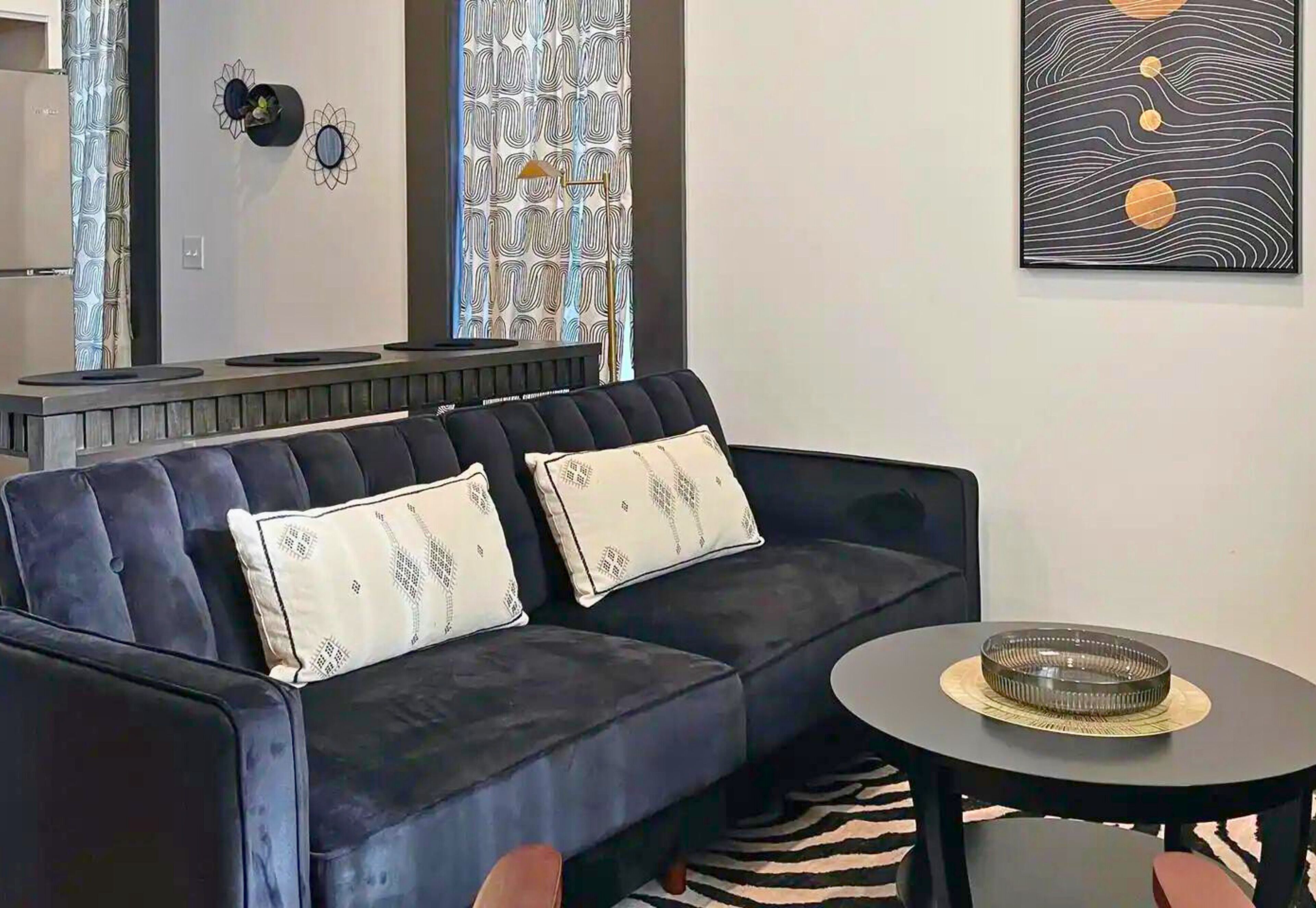 A modern living room with a dark blue sofa, two patterned pillows, a round coffee table with a decorative bowl, and a wall art piece.