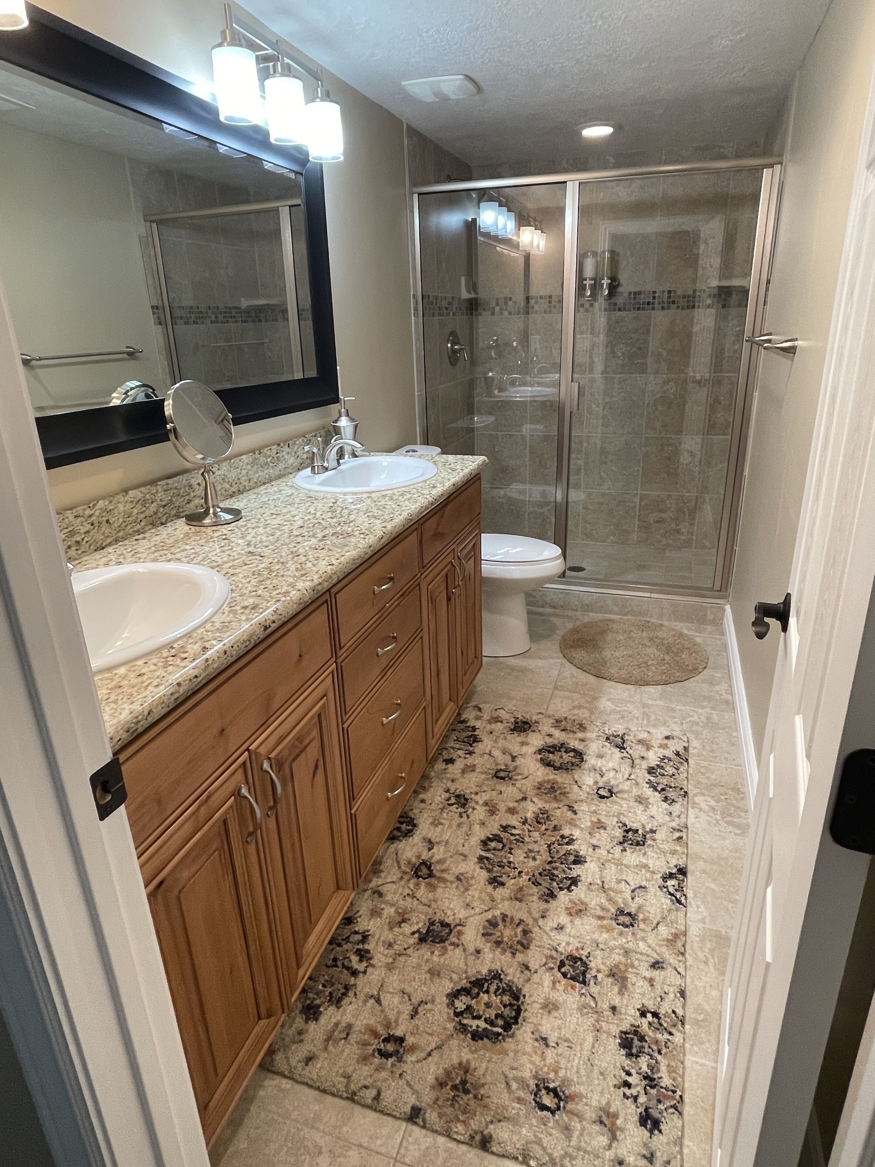 A modern bathroom with two sinks, a large mirror, a walk-in shower, and a patterned rug.