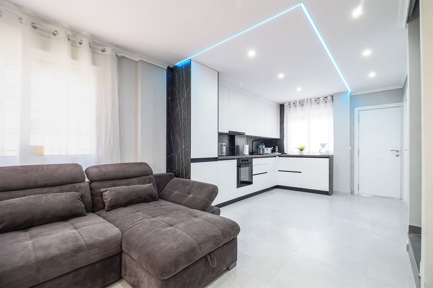 Een moderne woonkamer met een grijze modulaire bank, een keuken met witte kasten en zwarte aanrechtbladen, en een plafond met blauwe LED-verlichting.
