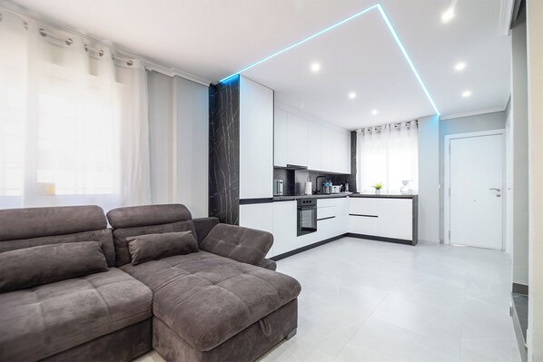 A modern living room with a grey sectional sofa, a kitchen with white cabinets and black countertops, and a ceiling with blue LED lighting. - Casa Olivia mit Beheizbaren Privatpool - 5 Min. vom Strand Entfernt (Torrevieja)