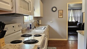 Une cuisine avec des meubles blancs, un four à micro-ondes, une cuisinière et un évier. Il y a une horloge au mur et un tapis sur le sol.