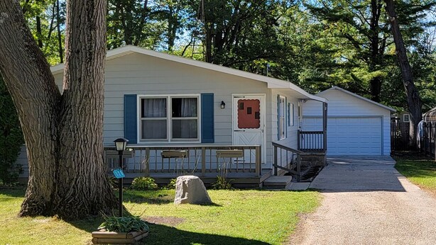Une maison mobile de plain-pied avec un porche à l’avant, un garage et un grand arbre au premier plan.