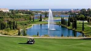 Ein Golfplatz mit Teich und Springbrunnen, umgeben von Bäumen und Gebäuden im Hintergrund.