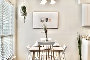 Uma sala de jantar com uma mesa posta para quatro pessoas, uma planta pendurada e uma obra de arte emoldurada na parede.