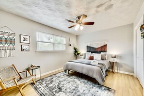 Quarto com cama, ventilador de teto, janela com persianas, planta suspensa na parede e tapete estampado.