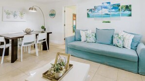 Een woonkamer met een turquoise bank, witte stoelen, een houten salontafel met decoratie, een plafondventilator en wanddecoratie.