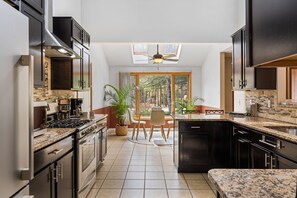A modern kitchen with dark cabinetry, a central island, and a dining area with a view of the forest.