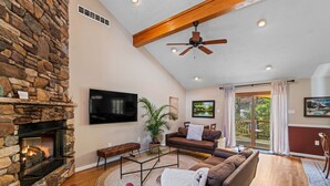 A spacious living room with a stone fireplace, a ceiling fan, a large flat-screen TV, a brown leather sofa, and a sliding glass door leading to a deck.