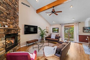 A spacious living room with a stone fireplace, a ceiling fan, a large flat-screen TV, a brown leather sofa, and a sliding glass door leading to a deck.