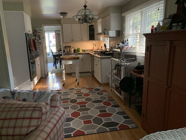A compact kitchen with white cabinets, a built-in oven, a microwave, and a gas stove. There's a wooden dining table with chairs, a patterned rug, and a window with blinds.