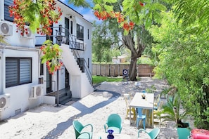 Un bâtiment blanc avec un escalier, un espace extérieur aménagé avec des chaises, et un arbre aux fleurs rouges.