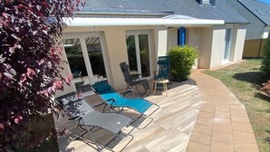 Un patio con terraza de madera curva, mobiliario exterior y una zona cubierta con puertas correderas de vidrio.