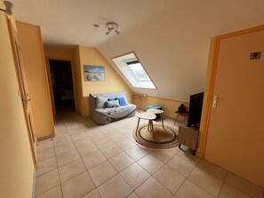 A compact living space with a sloped ceiling, a small seating area with a sofa and cushions, a television, and a door leading to another room.