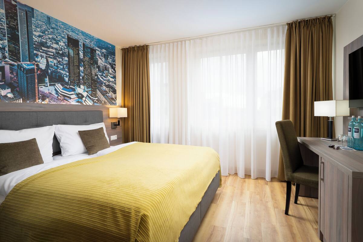 Une chambre d’hôtel avec un grand lit, un bureau, une chaise, une télévision et une fresque représentant un paysage urbain sur le mur.