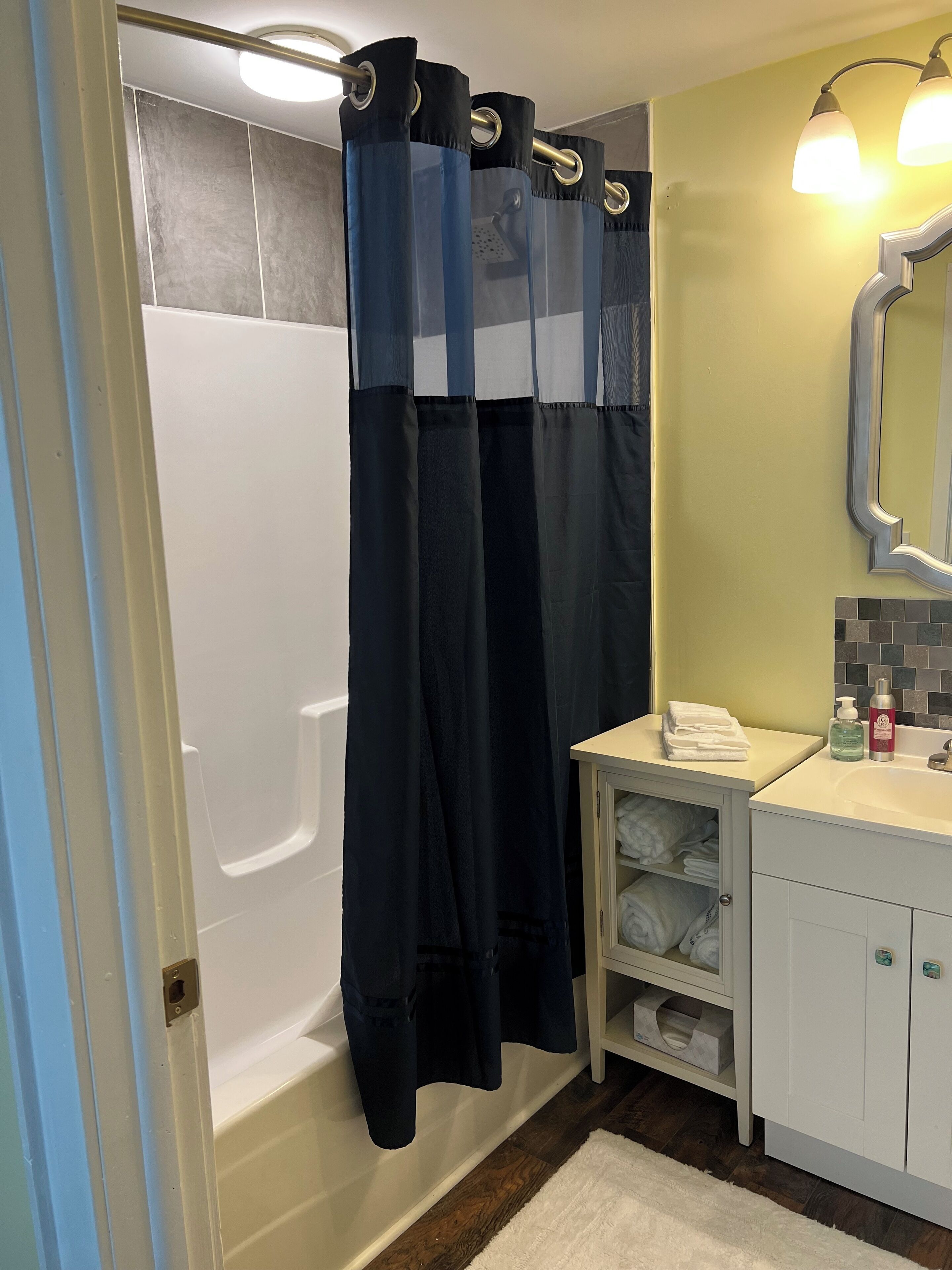 Un baño con bañera, cortina de ducha, lavabo con artículos de aseo y espejo.