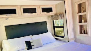 A bed with white linens and a black pillow, a headboard, and a window with curtains. There are wooden shelves above the bed. - Lakefront 1 bedroom tiny home (Acton)