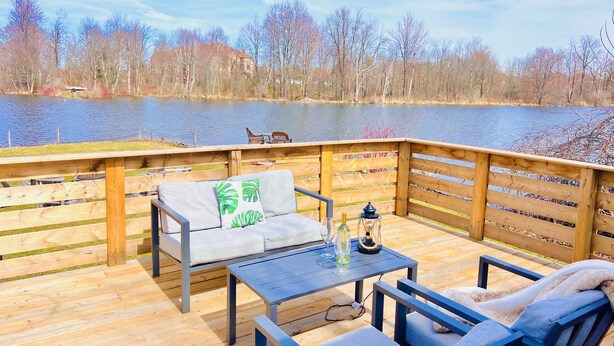 A wooden deck with outdoor furniture, a table, and a view of a lake and trees. - Lakefront 1 bedroom tiny home (Acton)