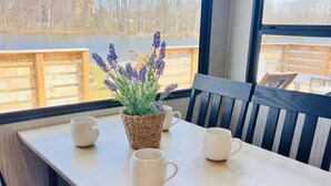 A table with a woven basket containing lavender, two white mugs, and a view of a lake and trees through large windows. - Lakefront 1 bedroom tiny home (Acton)