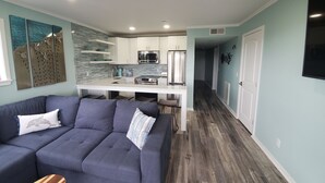 A modern living room with a dark blue sofa and a wooden coffee table, a kitchen with white cabinets and stainless steel appliances, and a hallway with wooden flooring. - Renovated Oceanview End Unit w/ Use of Hilton Ocean Oak Resort and Amenities (Hilton Head Island)