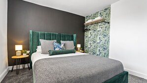 Un lit bien fait, avec une tête de lit recouverte d’un tissu vert, deux lampes de chevet et une petite table sur laquelle est posée une plante.